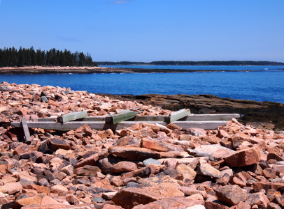 [Lots of large pinkish rocks on the shore with a grey wooden structure atop it that resembles fence posts connected with a top and bottom rail.]